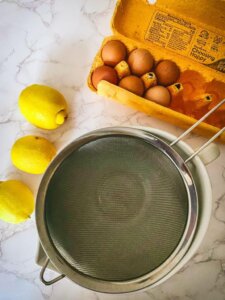 3 lemons next to an egg carton and bowl with a strainer over it